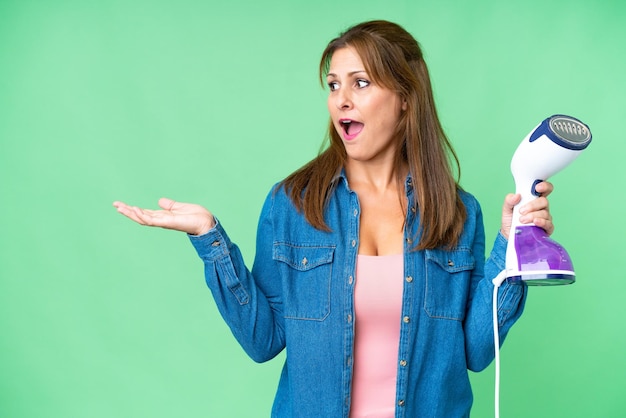 Mulher de meia-idade segurando um ferro sobre fundo isolado com expressão facial de surpresa