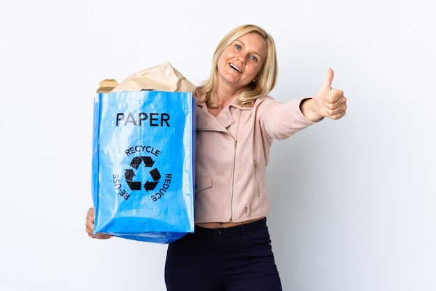 Mulher de meia-idade segurando um saco de reciclagem cheio de papel para reciclar isolado em uma parede branca com o polegar para cima porque algo bom aconteceu