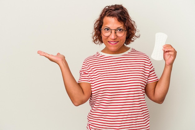 Mulher de meia idade segurando uma compressa isolada no fundo branco, mostrando um espaço de cópia na palma da mão e segurando a outra mão na cintura.