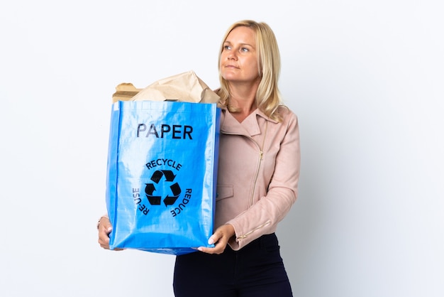 Mulher de meia-idade segurando uma sacola cheia de papel para reciclar isolada em uma parede branca olhando para o lado