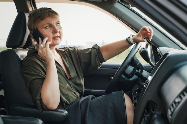 Mulher de meia idade sentada ao volante de um carro e fala indignada ao telefone