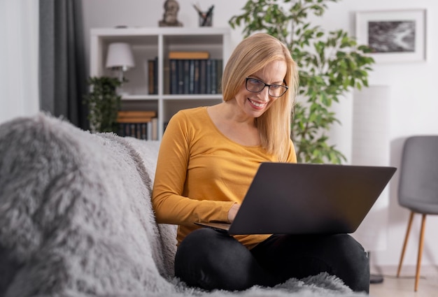 Mulher de meia idade sentada no sofá com laptop moderno de joelhos e digitando no teclado Senhora caucasiana com cabelos loiros usando óculos e roupas casuais