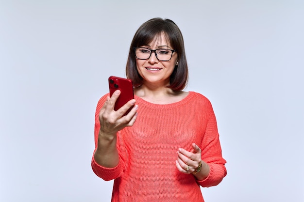 Mulher de meia-idade sorridente olhando para o smartphone em um fundo de estúdio leve Mulher bonita e emocional usando aplicativos móveis on-line Tecnologia estilo de vida internet conceito de pessoas de 40 anos de idade