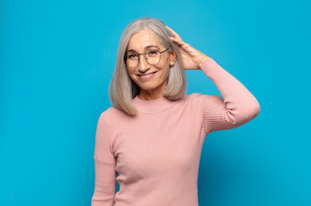 Mulher de meia-idade sorrindo alegre e casualmente, levando a mão na cabeça com um olhar positivo, feliz e confiante