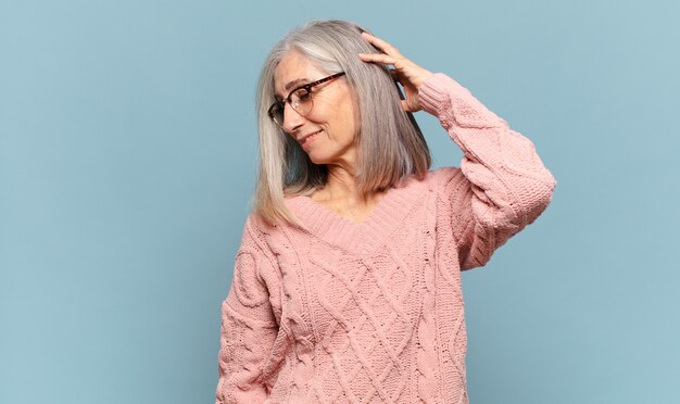 Mulher de meia-idade sorrindo alegre e casualmente, levando a mão na cabeça com um olhar positivo, feliz e confiante