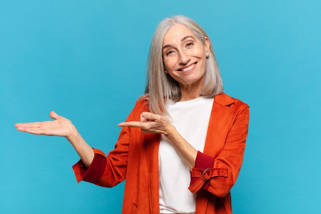 Mulher de meia-idade sorrindo alegremente e apontando para copiar o espaço na palma da mão ao lado, mostrando ou anunciando um objeto