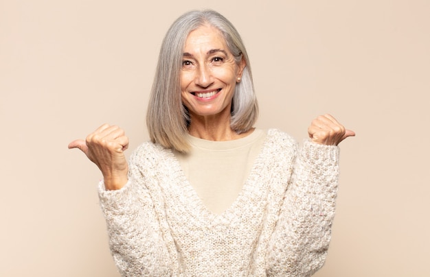 Mulher de meia idade sorrindo alegremente e parecendo feliz, sentindo-se despreocupada e positiva com os dois polegares para cima