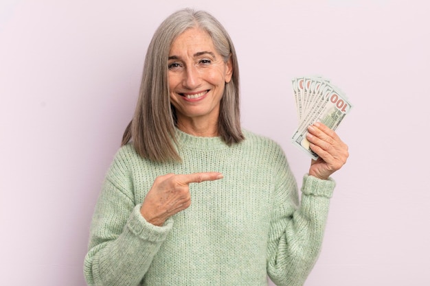Mulher de meia idade sorrindo alegremente se sentindo feliz e apontando para o conceito de notas de dólar lateral
