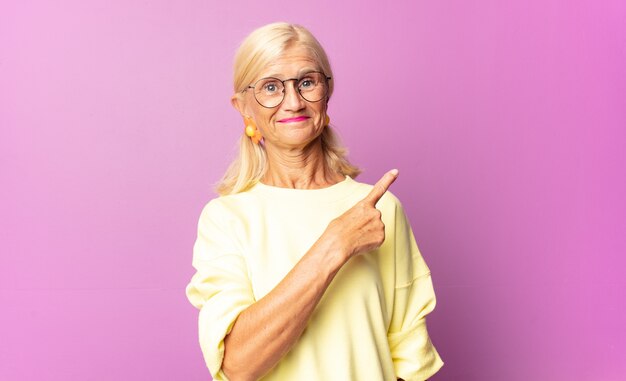 Mulher de meia-idade sorrindo alegremente, sentindo-se feliz e apontando para o lado e para cima, mostrando o objeto no espaço da cópia