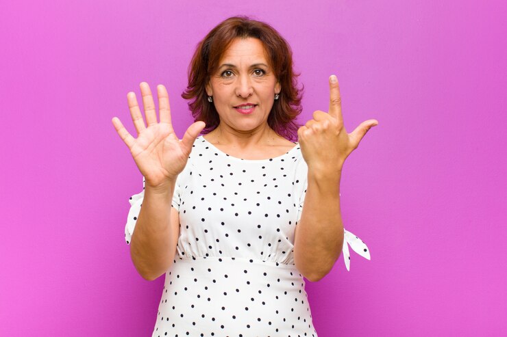 Foto mulher de meia idade sorrindo e olhando amigável, mostrando o número sete ou sétimo com a mão para a frente, contagem regressiva contra a parede roxa