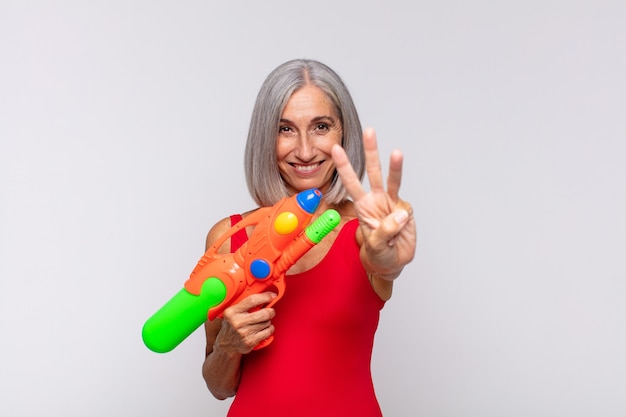 Mulher de meia idade sorrindo e parecendo amigável, mostrando o número três ou o terceiro com a mão para a frente, fazendo a contagem regressiva com uma pistola d'água