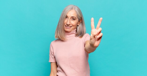 Mulher de meia-idade sorrindo e parecendo feliz, despreocupada e positiva, gesticulando vitória ou paz com uma das mãos