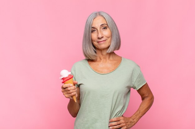 Mulher de meia idade sorrindo feliz com uma mão no quadril e uma atitude confiante, positiva, orgulhosa e amigável tomando um sorvete