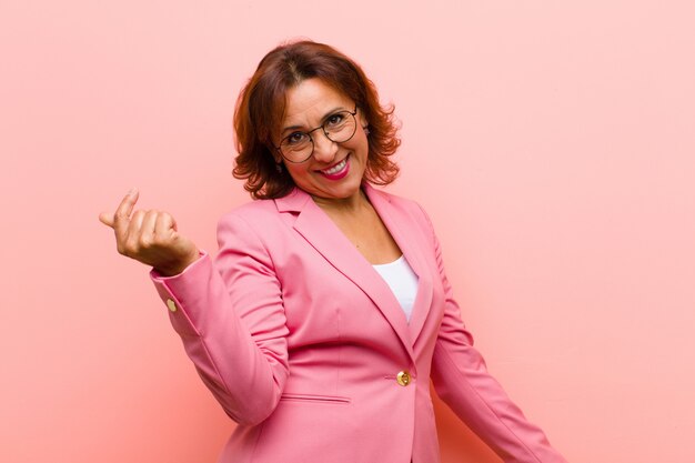 Mulher de meia idade sorrindo, sentindo-se despreocupada, relaxada e feliz, dançando e ouvindo música, se divertindo em uma festa contra a parede rosa