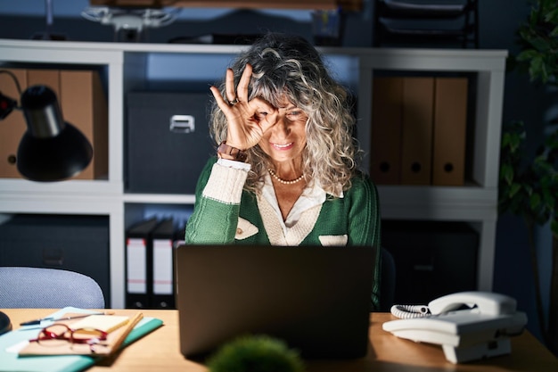 Mulher de meia-idade trabalhando à noite usando computador portátil fazendo gesto ok com a mão sorrindo olho olhando através dos dedos com rosto feliz