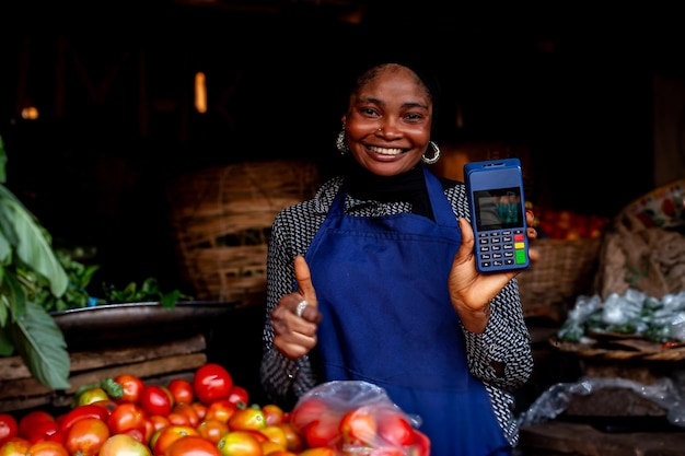 Mulher de mercado nigeriana feliz dando o polegar para cima segurando uma máquina de pagamento vendendo tomates