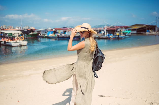 Mulher de mochileiro loira viajante na vista traseira do chapéu de palha caminha ao longo da costa nos barcos de pesca do cais. Aventura de viagens no turista de China.Tropical ilha Ásia. Conceito de viagem de férias de férias de verão