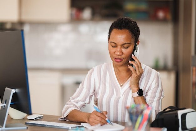 Mulher de negócios africana falando em um smartphone enquanto faz anotações em seu escritório em casa.