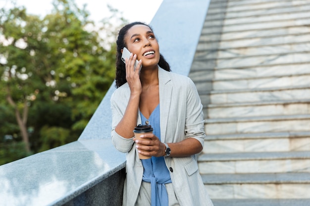 Mulher de negócios africana sorridente atraente em pé ao ar livre na cidade, encostada na escada, segurando uma xícara de café para viagem, falando no celular