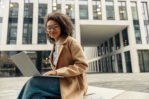 Mulher de negócios africana sorridente trabalhando laptop enquanto está sentada do lado de fora no fundo do escritório