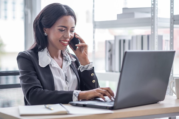 Mulher de negócios africana trabalhando no escritório usando conversa de computador por telefone inteligente com cliente