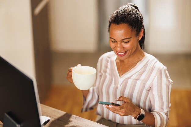 Mulher de negócios africana usando um celular e tomando café enquanto trabalha em casa.