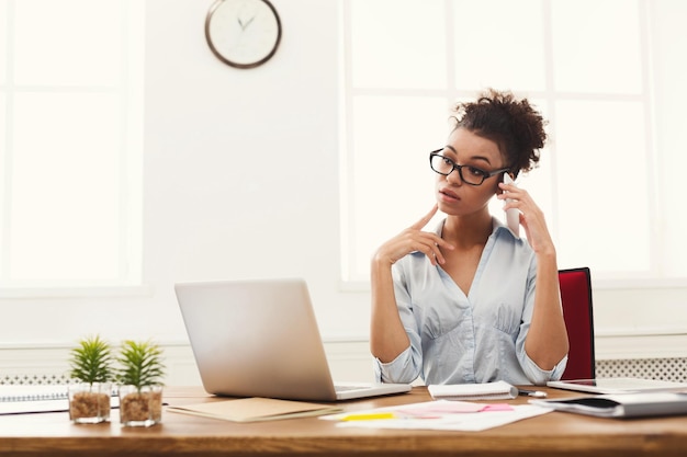 Mulher de negócios afro-americana atenciosa no trabalho, falando no telefone, sentada em seu local de trabalho no escritório, copie o espaço