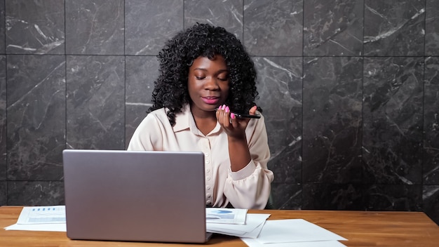 Mulher de negócios afro-americana com cabelo encaracolado fala no smartphone e olha gráficos em papéis, sentada na mesa com o laptop contra a parede de mármore