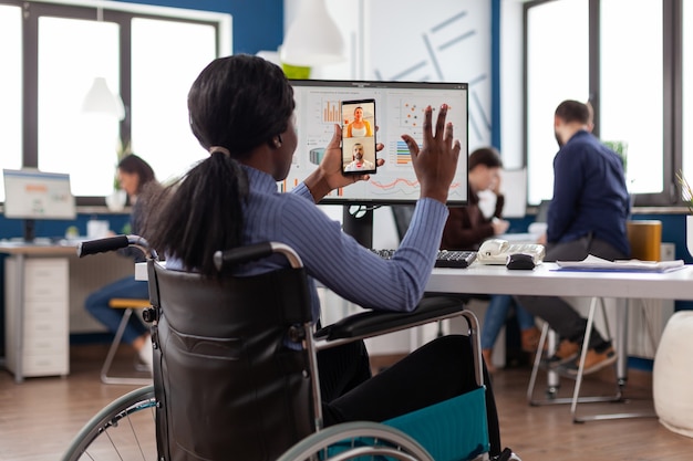 Mulher de negócios afro-americana com deficiência e deficiência cumprimentando o trabalho em equipe remoto durante a conferência de videochamada online no escritório de negócios de inicialização. Chamada de teleconferência na tela do telefone