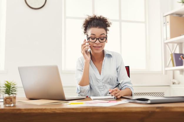 Mulher de negócios afro-americana concentrada no trabalho, falando no telefone, sentada em seu local de trabalho no escritório, copie o espaço