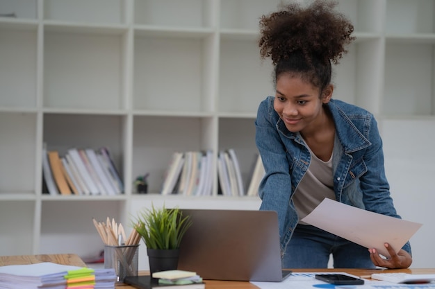 Mulher de negócios afro-americana em uma jaqueta de jeans de pé e usando um computador portátil em um escritório
