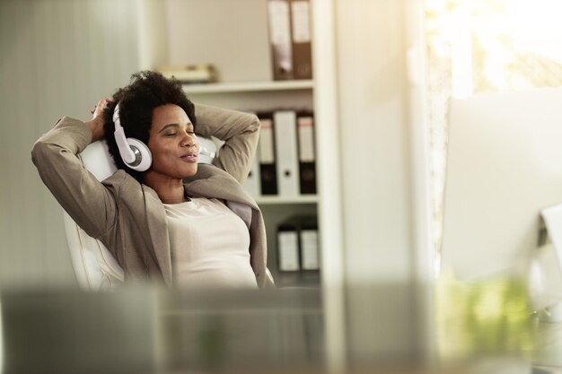 Mulher de negócios afro-americana relaxando e ouvindo música nos fones de ouvido enquanto desfruta de uma pausa para esticar com as mãos atrás da cabeça em um escritório sozinha no trabalho.