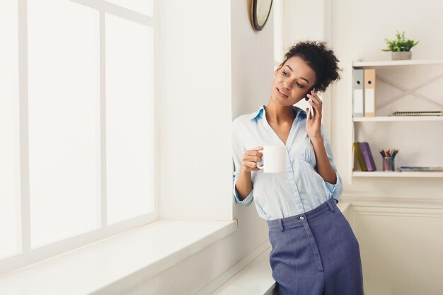 Foto mulher de negócios afro-americana sorridente, falando por telefone perto da janela no escritório, bebendo café. comunicação, tecnologia, conceito de sucesso