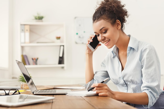 Foto mulher de negócios afro-americana sorridente no trabalho, falando no telefone e fazendo anotações, sentada em seu local de trabalho no escritório, copie o espaço