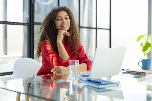 Mulher de negócios alegre atraente na camisa vermelha trabalhando no laptop no escritório moderno
