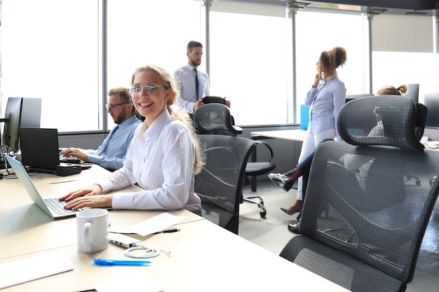 Mulher de negócios alegre em trajes formais, trabalhando no laptop no escritório moderno com colegas no fundo.