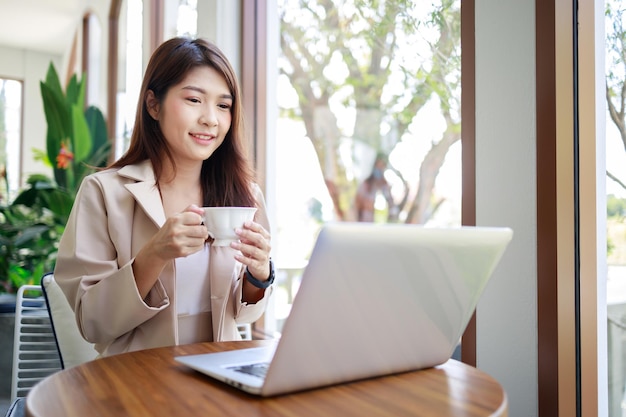 Mulher de negócios asiática atraente bebendo café e trabalhando por laptop na cafeteria Mulher confiante e inteligente e conceito de negócios