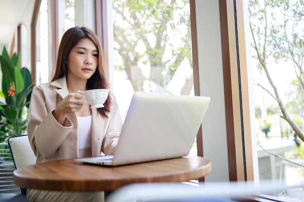 Mulher de negócios asiática atraente bebendo café e trabalhando por laptop na cafeteria