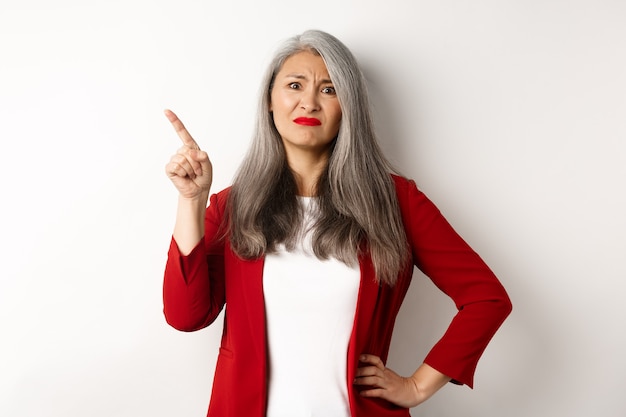 Mulher de negócios asiática cética usando um blazer vermelho fazendo careta, apontando para o canto superior esquerdo e parecendo insatisfeita, desaprova algo, parada sobre um fundo branco