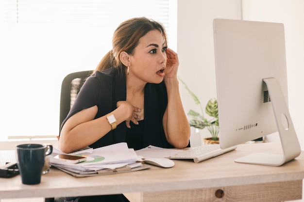 Mulher de negócios asiática chocada sentada em uma mesa e olhando para as notícias na tela do computador