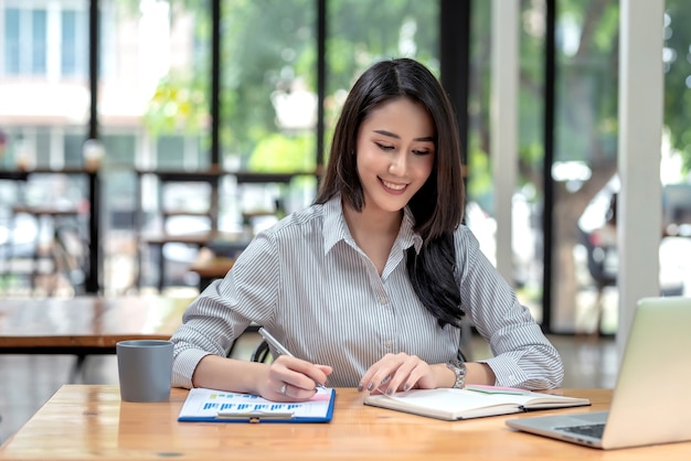 Mulher de negócios asiática encantadora sentado no escritório segurando caneta documento escrevendo notebook laptop no trabalho estatístico de mesa.