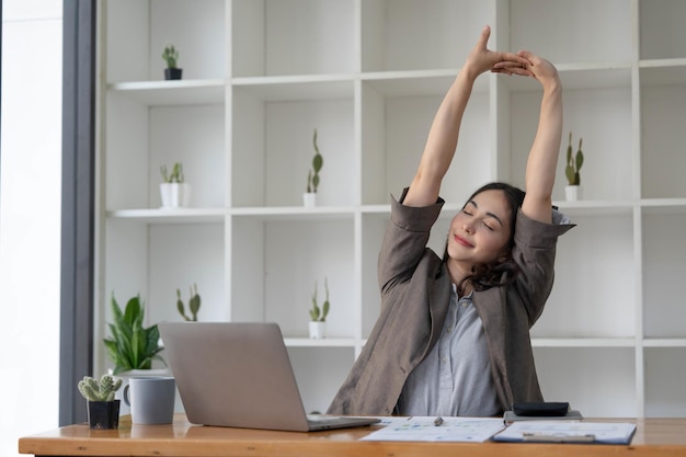 Mulher de negócios asiática estende os braços para relaxar seus músculos cansados de trabalhar em sua mesa o dia todo no escritório