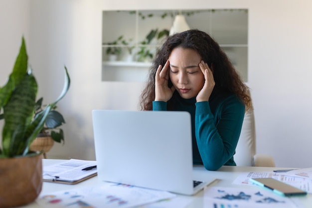 Mulher de negócios asiática exausta com dor de cabeça no escritório Mulher criativa trabalhando na mesa do escritório sentindo-se cansada Mulher de negócio casual estressada sentindo dor de cabeça enquanto trabalha demais no computador portátil