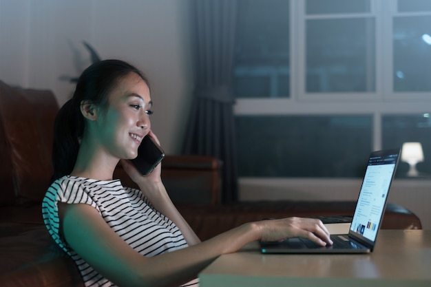 Foto mulher de negócios asiática falando no smartphone trabalhando no laptop, hora extra, tarde da noite em casa