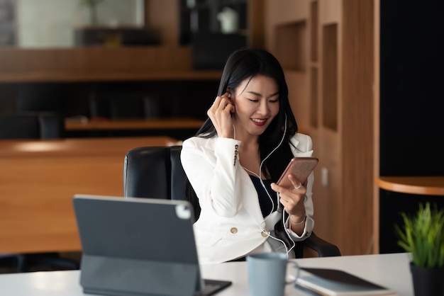 Mulher de negócios asiática feliz sorrindo usando fones de ouvido e usando um smartphone no escritório.