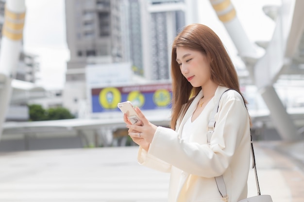 Mulher de negócios asiática jovem bonita está indo para o escritório ou local de trabalho, que tem o smartphone nas mãos em uma cidade grande, com edifícios comerciais com a cidade como pano de fundo.