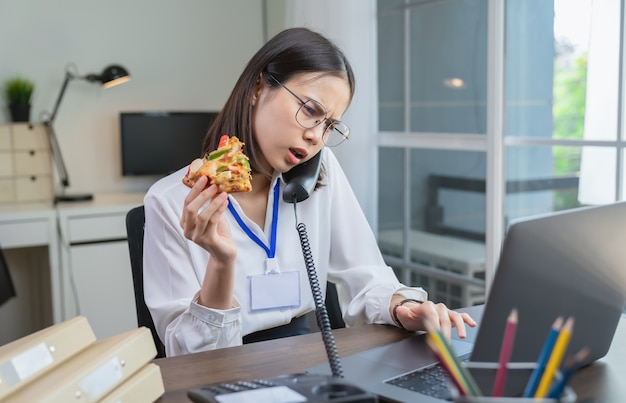 Foto mulher de negócios asiática nova que come o petisco da pizza ao falar o telefone com os clientes devido ao trabalho tarde.