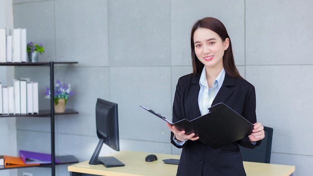 Mulher de negócios asiática profissional de terno preto confiante sorri alegremente enquanto trabalha e segura