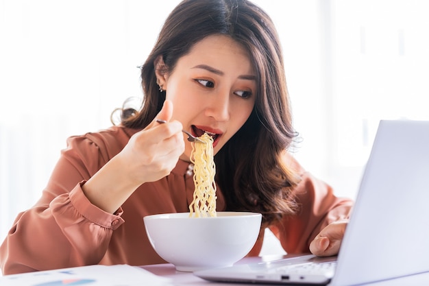 Mulher de negócios asiática tem que comer macarrão enquanto trabalha