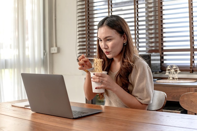 Mulher de negócios asiática trabalhando sozinha online em casa. Mulher comendo macarrão de xícara e ocupado.